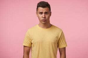 Bewildered young handsome short haired brunette man grimacing his face while looking confusedly at camera, keeping hands down while posing over pink background photo