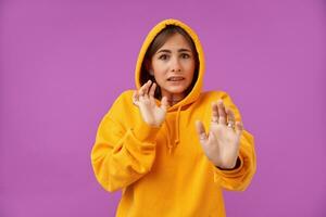 Young pretty woman with shocked face, showing sign of worried, defense. Frightened, looking at the camera over purple background. Wearing orange hoodie, teeth braces and rings photo