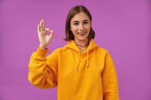 Female student, young lady with short brunette hair. Shows a sign that all is okay. Satisfied girl look at the camera over purple background. Wearing orange hoodie, teeth braces and rings photo