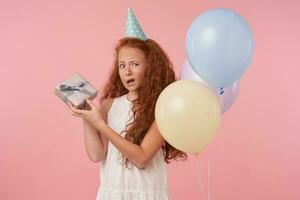 Portrait of curly female kid with long foxy hair holding gift-wrapped box and looking in camera with pout, standing over pink background with colored air balloons photo