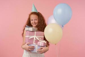 Portrait of cute little girl redhead with foxy curly hair in elegant dress and birthday cap happily looking in camera with gift box in hands over pink background. Children and celebration concept photo