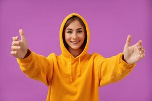 Portrait of attractive, nice looking, happy girl with big smile, shows that she wants a hug. Look at the camera over purple background. Wearing orange hoodie, teeth braces and rings photo