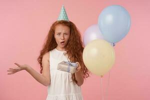 Horizontal shot of redhead littl girl with long curly hair standing over pink background in white elegant dress and birthday cap, holding present in hand and looking in camera with unpleased face photo