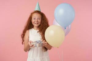 Studio shot of joyful redhead little girl with long curly hair holding gift-wrapped box being excited and surprised to get birthday present, standing over pink background in festive clothes photo