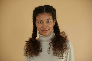 Portrait of attractive positive young curly brunette woman with dark skin dressed in roll-neck woolen sweater while standing over beige background, looking to camera with folded lips photo