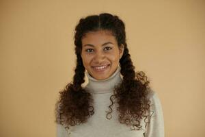 Close-up of lovel young curly dark skinned female with braids having freckles on her face and wearing natural make up while posing over beige background in woolen sweater photo