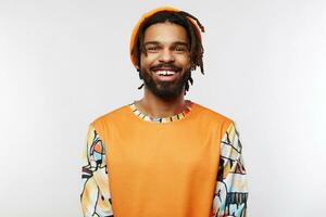 Happy young dark skinned brunette male with dreadlocks showing his white perfect teeth while looking cheerfully at camera, standing over white background with hands down photo