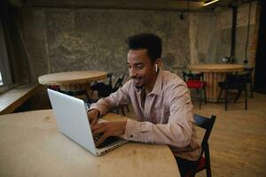 Indoor shot of young dark skinned man with beard wearing casual clothes while sitting at table with laptop and typing text on keyboard, posing over city cafe interior, looking at screen and smiling photo