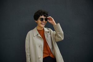 Fashionable young curly woman with short haircut wearing trendy outfit while walking outside, posing over black urban wall, keeping hand on her sunglasses and smiling cheerfully photo