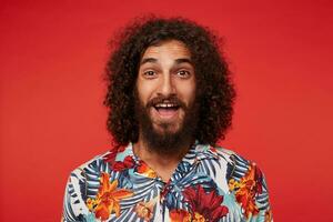 Indoor shot of happy young brunette bearded man with curls looking joyfully to camera with wide smile, being in high spirit while posing over red background, dressed in multi-colored flowered shirt photo