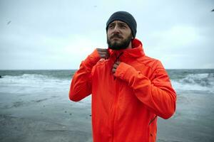 Sporty serious looking young dark haired bearded man with eyebrow piercing wearing black cap and winter sporty coat with hood, posing over coastline of sea on cold stormy weather photo