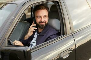 retrato de joven hermoso exitoso barbado hombre en un azul chaqueta y a rayas camiseta, se sienta detrás el rueda de el coche y murga para un respuesta en el telefon desde su novia. foto