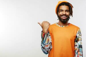 Cheerful young dark skinned brunette man showing aside with thumb and looking positively at camera while standing over white background in orange hat and casual pullover photo