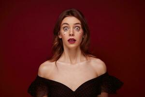 Open-eyed young pretty female with brown wavy hair wearing festive evening makeup while standing over claret background, rounding amazedly eyes and looking at camera with shocked face photo