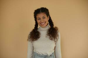 Indoor shot of pretty curly dark skinned brunette lady with braids looking on camera with charming smile while standing over beige background, keeping hands along body photo