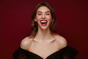 Cheerful attractive young brunette lady with festive makeup dressed in elegant top laughing happily with wide smile while standing against claret background, being in high spirit photo