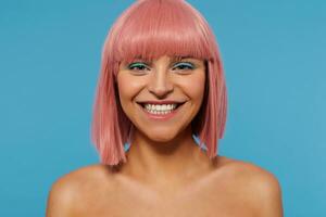 Close-up of happy young attractive pink haired female with colored makeup showing her perfect white teeth while looking cheerfully at camera, isolated over blue background photo