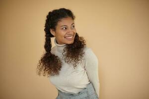 Sudio photo of attractive young dark sinned female with curly long hair being braided, looking aside happily and keeping hands behind her back, isolated over beige background