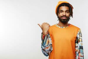 Positive young pretty dark skinned male with dreadlocks keeping his lips folded while pointing aside with thumb, wearing orange hat and pullover while posing over white background photo