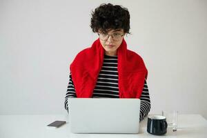 Serious young dark haired curly woman with short trendy haircut wearing eyeglasses while working over white background with modern laptop, looking at screen with concentrated face photo