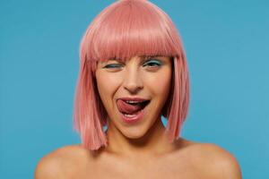 Coquettish young pretty woman with short pink hair giving wink while looking playfully at camera, sticking out her tongue while standing over blue background photo