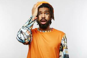 Confused young brown-eyed dark skinned male with beard raising emotionally hand to his head and looking at camera with puzzled face, standing over white background photo