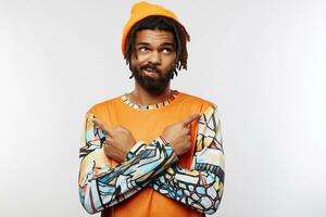 Displeased young handsome dark skinned guy with beard twisting his mouth while looking upwards and pointing in different sides with raised forefingers, isolated over white background photo