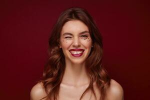Positive young attractive brunette woman with wavy hairstyle wearing evening makeup while posing over claret background, looking cheerfully at camera and winking with one eye photo
