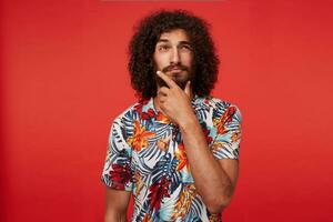 Pensive handsome young brunette curly male with beard holding his chin with raised hand and looking thoughtfully aside, squinting and wrinkling his forehead while standing over red background photo