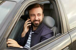 Photo of of young attractive successful bearded man in a blue jacket and striped t-shirt, sits behind the wheel of the car and waits for a response on the telefon.