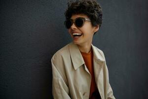 Close-up of young curly fashionable female with casual hairstyle posing over city black wall, wearing stylish clothes and sunglasses, looking aside cheerfully with broad sincere smile photo