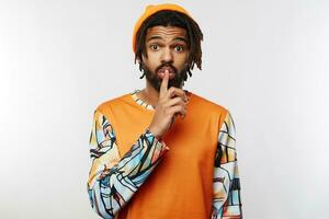 Indoor photo of young dark skinned brunette man with beard raising hand with hush gesture and looking surprisedly at camera, standing over white background