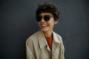 Outdoor portrait of dark haired young curly woman wearing trendy outfit and stylish sunglasses, smiling widely and showing her pleasant emotions while posing over city environment photo