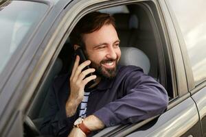 Young smiling attractive successful bearded man in a blue jacket and striped t-shirt, sits behind the wheel of the car in traffic, so as not to be bored called his friend and tells funny stories. photo