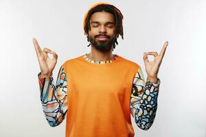 Pleasant looking young unshaved brunette guy with dark skin folding hands in mudra gesture and keeping his eyes closed while standing over white background photo