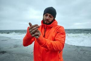 Image of attractive young athletic brunette male with beard standing over seaside on grey stormy weather and wearing warm sporty clothes, preparing for morning workout photo