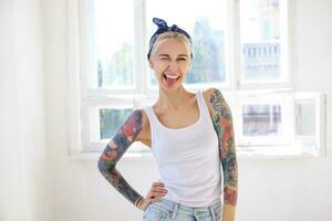 Joyful young attractive tattooed lady with casual hairstyle giving cheerfully wink at camera and showing tongue, keeping raised hand on waist while posing over bright studio photo