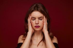 Stressed young brunette lady with curls looking tired and holding her face with hands, keeping eyes closed and frowning eyebrows while posing over claret background photo