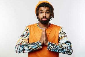 Confused young pretty bearded brunette guy with dreadlocks wrinkling forehead while looking at camera and folding raised hands, isolated over white background photo