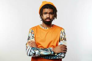 Unhappy young dark skinned brunette man crossing his arms while posing over white background, frowning his eyebrows while looking sadly at camera with folded lips photo