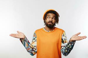 Confused young bearded brunette male with dreadlocks raising emotionally palms and looking perplexedly upwards, standing over white background in casual wear photo