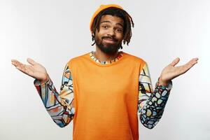 Bewildered young pretty dark skinned brunette man dressed in orange hat and pullover keeping lips folded while shrugging with raised palms, standing over white background photo