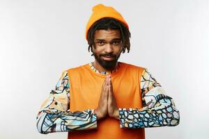 Glad young handsome dark skinned male with beard keeping raised palms together while looking positively at camera with light smile, isolated over white background photo