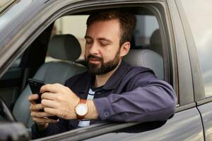 Young hendsome successful bearded man in a blue jacket and striped t-shirt, sits behind the wheel of the car, chatting with colleague by phone. photo