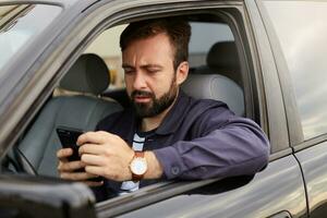 Portrait of frowning dosconected bearded man in a blue jacket and striped t-shirt, sits behind the wheel of the car, chatting with colleague by phone, displeased with the argument. photo