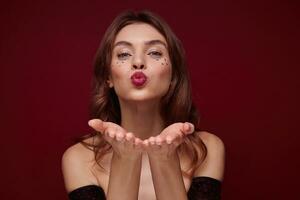 Close-up of cheerful pretty young brown haired woman with festive makeup keeping palms raised while folding lips for air kiss, looking positively at camera while posing over claret background photo