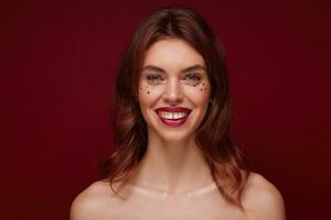 Close-up of happy pretty young brunette woman with evening makeup looking cheerfully to camera with charming smile, showing her white perfect teeth while standing over claret background photo