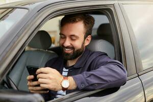 Portrait of young hendsome successful bearded man in a blue jacket and striped t-shirt, sits behind the wheel of the car, chatting with colleague by phone, broadly smiling, saw a funny picture photo