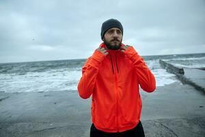 Pensive young sporty bearded male in black cap and warm orange coat looking seriously ahead and keeping lips folded, going to make morning exercises by sea. Fitness male model photo
