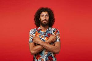 Surprised young brunette curly man with beard keeping crossed hands on his chest and looking to camera with wide eyes opened, standing over red background in casual clothes photo